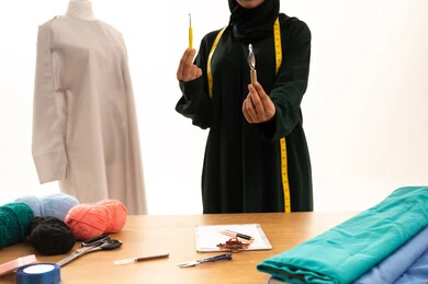 New and modern designs, the importance of women in the field of sewing, a portrait of an Arab Gulf Saudi woman wearing an abaya holding a crochet needle in her hand, a fashion design workshop, women's professions and artistic works, the concept of the world of fashion and style, white background.