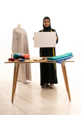 A blank billboard, looking at the camera with gestures of joy and satisfaction, a white square mockup, a portrait of a Saudi Gulf Arab seamstress wearing a hijab and a black abaya, smiling while holding a white square board, crafts and manual professions, a fashion design workshop, sewing skills and mastery of women's clothing tailoring, full-body shot, white background.