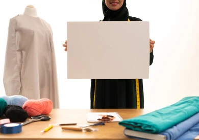 ورشة عمل تصميم ازياء، موك اب مربع أبيض، لوح إعلاني فارغ، بورتريه لخياطة عربية خليجية سعودية ترتدي الحجاب و العباءة السوداء مبتسمة تحمل لوحه بيضاء مربعه، حرف ومهن يدوية، مهارة الخياطة واتقان تفصيل الملابس النسائية، خلفية بيضاء