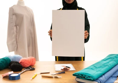 موك اب مربع أبيض، لوح إعلاني فارغ، بورتريه لخياطة عربية خليجية سعودية ترتدي الحجاب و العباءة السوداء مبتسمة تحمل لوحه بيضاء مربعه، حرف ومهن يدوية، ورشة عمل تصميم ازياء، مهارة الخياطة واتقان تفصيل الملابس النسائية، خلفية بيضاء