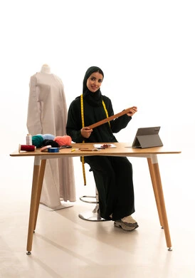 حرف ومهن يدوية، استعمال أدوات ومعدات الخياطة، إتقان صنع الملابس السعودية، بورتريه لخياطة عربية خليجية سعودية ترتدي الحجاب و العباءة السوداء تمسك بيديها المسطرة الخشبية ، ايماءات السعادة و السرور، خلفية بيضاء