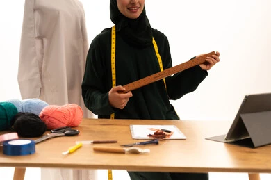 Using sewing tools and equipment, mastering the making of Saudi clothing, crafts and manual professions, a close-up portrait of a Saudi Gulf Arab seamstress wearing a hijab and a black abaya holding a wooden ruler in her hands, gestures of happiness and joy, white background.
