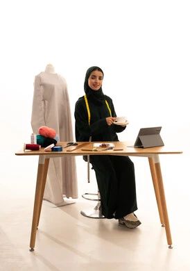 تناول المشروبات الساخنة اللذيذة بإيماءات السرور، ورشة عمل تصميم ازياء، مهن وأعمال فن الخياطة، بورترية لمصممة أزياء عربية خليجية سعودية ترتدي الحجاب و عباءة سوداء تحمل فنجان من القهوه ، أدوات الخياطة، مهارة الخياطة واتقان تفصيل الملابس النسائية، الأزياء والموضة، خلفية بيضاء