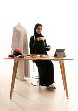 Enjoying hot beverages, a fashion design workshop, professions and crafts of sewing art, a portrait of an Arab Gulf Saudi fashion designer wearing a hijab and a black abaya holding a cup of coffee, sewing tools, sewing skills and mastery of women's clothing tailoring, fashion and style, white background.