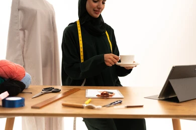 Spending time sipping a favorite drink, a fashion design workshop, professions and crafts of sewing art, a portrait of an Arab Gulf Saudi fashion designer wearing a hijab and a black abaya holding a cup of coffee, sewing tools, sewing skills and mastery of women's clothing tailoring, fashion and style, white background.