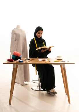 Fashion design workshop, professions and crafts of sewing art, a portrait of an Arab Gulf Saudi fashion designer wearing a hijab and a black abaya holding a book and reading something, sewing tools, sewing skills and mastery of women's clothing tailoring, fashion and style, the concept of self-education, white background.