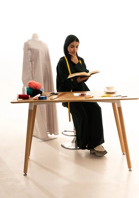 التمعن والتركيز والتخطيط للمستقبل، مهن وأعمال فن الخياطة، بورترية لمصممة أزياء عربية خليجية سعودية ترتدي الحجاب و عباءة سوداء تحمل كتاب تضع يدها على ذقنها بايماءات التفكير والتأمل، ورشة عمل تصميم ازياء، أدوات الخياطة، مهارة الخياطة واتقان تفصيل الملابس النسائية