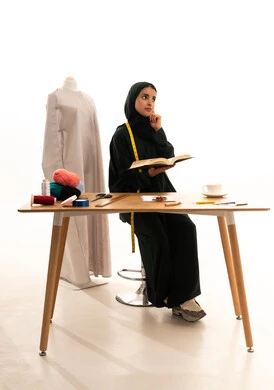 Trying to remember something, professions and works of the art of sewing, a portrait of an Arab Gulf Saudi fashion designer wearing a hijab and a black abaya holding a book in her hand, a fashion design workshop, sewing tools, sewing skills, and mastery of women's clothing tailoring.