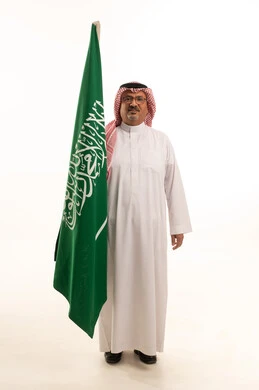 ذكرى اليوم الوطني السعودي، التقدم بالعمر والهيبة، بورتريه  لرجل كبير بالسن عربي خليجي سعودي يرتدي الثوب التقليدي و الشماغ يقف باستقامة ويمسك علم المملكة, الاهتمام المظهر الخارجي، مفهوم الرجولة العربية, الجسم بالطول الكامل، خلفية بيضاء