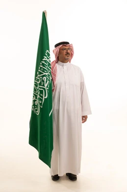 Celebrating national identity, the anniversary of the Saudi National Day, aging and dignity, a side portrait of an elderly Arab Gulf Saudi man wearing traditional attire and a shemagh standing upright holding the Kingdom's flag, the concept of Arab masculinity, full-body, white background.