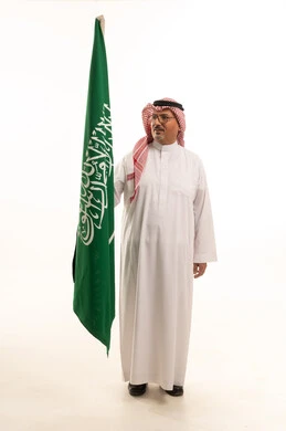 الاحتفال بالعيد الوطني السعودي، التقدم بالعمر والهيبة، بورتريه  لرجل كبير بالسن عربي خليجي سعودي يرتدي الثوب التقليدي و الشماغ يقف باستقامة ويمسك علم المملكة, الاهتمام المظهر الخارجي، مفهوم الرجولة العربية, الجسم بالطول الكامل، خلفية بيضاء