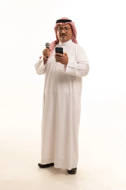 Integrating technology with training on singing talents, presenting cultural segments, skill and art of delivery, a portrait of an elderly Arab Gulf Saudi man wearing traditional attire and a ghutrah standing upright holding a microphone and a mobile phone, the concept of Arab masculinity, full-body, white background.