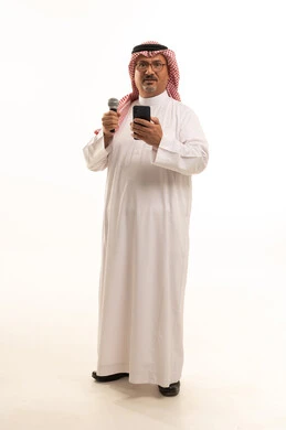 استخدام الهاتف المحمول مع مهارة وفن الألقاء، تقديم فقرات ثقافية، بورتريه من لرجل كبير بالسن عربي خليجي سعودي يرتدي الثوب التقليدي و الشماغ يقف باستقامة و يحمل بيده الميكروفون وهاتف محمول, مفهوم الرجولة العربية, الجسم بالطول الكامل، خلفية بيضاء