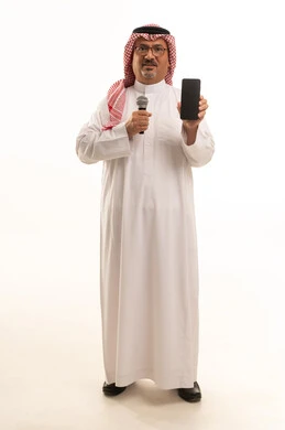 A black screen on the mobile phone, with skill and art of presentation, presenting cultural segments, a portrait of an elderly Arab Gulf Saudi man wearing traditional attire and a ghutrah standing upright holding a microphone and a mobile phone, the concept of Arab masculinity, full-body shot, white background.