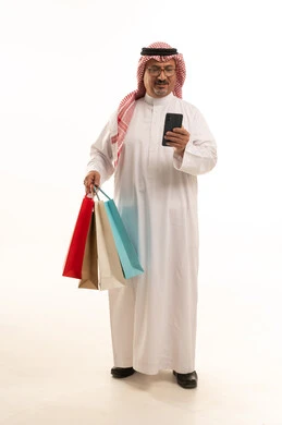 التسوق عبر الانترنت، الاستفادة من الحسومات الموسمية، إيماءات السعادة والسرور بالخصومات، بورتريه لرجل كبير بالسن عربي خليجي سعودي يرتدي الشماغ و الثوب التقليدي يحمل بيده الاكياس الورقية وهاتف، التسوق والحصول على العروض، صورة الجسم بالطول الكامل، خلفية بيضاء
