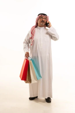 استخدام الانترنت في مجال التسوق، الاستفادة من الحسومات الموسمية، إيماءات السعادة والسرور، بورتريه لرجل كبير بالسن عربي خليجي سعودي يرتدي الشماغ و الثوب التقليدي يحمل بيده الاكياس الورقية والهاتف، التسوق والحصول على العروض، صورة الجسم بالطول الكامل، خلفية بيضاء