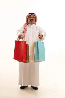 الاستفادة من الحسومات الموسمية، إيماءات السعادة والسرور بالخصومات، بورتريه لرجل كبير بالسن عربي خليجي سعودي يرتدي الشماغ و الثوب التقليدي يحمل بيده الاكياس الورقية الملونة، التسوق والحصول على العروض، صورة الجسم بالطول الكامل، خلفية بيضاء