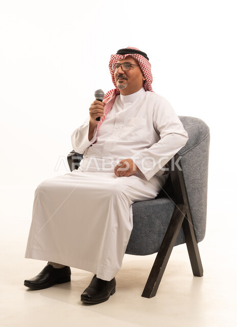 تعابير الاندماج والتفاعل في بودكاست، تسجيل مقطع صوتي غنائي، بورتريه لرجل مسن عربي خليجي سعودي يرتدي الثوب التقليدي والشماغ يجلس على كرسي مريح يمسك بيده المايكروفون، إيماءات الحماس والسرور، تقديم العروض الصوتية، ممارسة موهبة الغناء، خلفية بيضاء