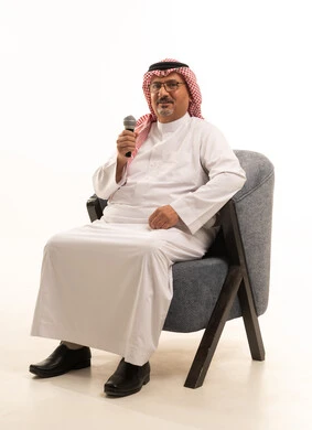 النظر إلى الكاميرا بإيماءات السرور، تعابير الاندماج والتفاعل، تسجيل مقطع صوتي غنائي، بورتريه لرجل مسن عربي خليجي سعودي يرتدي الثوب التقليدي والشماغ يجلس على كرسي مريح يمسك بيده المايكروفون، تقديم العروض الصوتية، ممارسة موهبة الغناء، خلفية بيضاء
