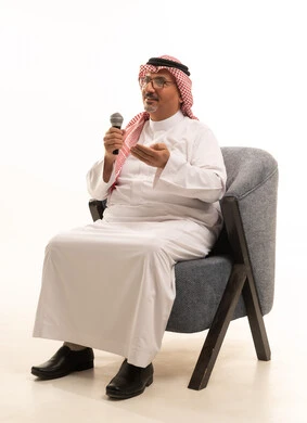 النظر إلى الكاميرا بإيماءات السرور، تعابير الاندماج والتفاعل، تسجيل مقطع صوتي غنائي، بورتريه لرجل مسن عربي خليجي سعودي يرتدي الثوب التقليدي والشماغ يجلس على كرسي مريح يمسك بيده المايكروفون، تقديم العروض الصوتية، ممارسة موهبة الغناء، خلفية بيضاء