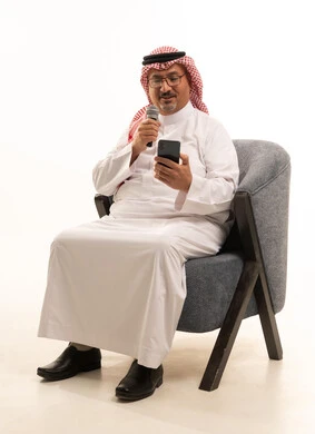 Reading from the phone while presenting a program, expressions of integration and interaction, recording a singing audio clip, a portrait of an elderly Arab Gulf Saudi man wearing traditional attire and a ghutrah sitting on a comfortable chair holding a microphone, presenting audio performances, practicing singing talent, white background.