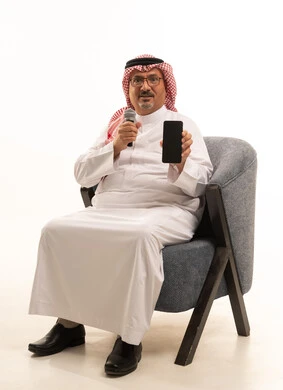 A black screen on the phone, expressions of engagement and interaction, recording a singing audio clip, a portrait of an elderly Arab Gulf Saudi man wearing traditional attire and a ghutrah sitting on a comfortable chair holding a microphone, presenting audio performances, practicing the talent of singing, white background.