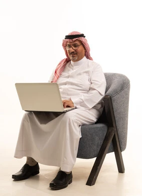 الجلوس على كرسي مريح واستخدام التقنيات الحديثة في العمل عن بعد، انهاء المهام اليومية، بورتريه مقرب لرجل كبير السن عربي خليجي سعودي يرتدي الزي التقليدي والشماغ يعمل عبر اللابتوب، إنجاز المهام اونلاين من خلال جهاز الحاسوب، خلفية بيضاء .