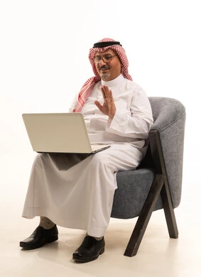 Making a video call on a mobile device, using modern technologies for remote work, completing daily tasks, a close-up portrait of an elderly Arab Gulf Saudi man wearing traditional attire and a ghutrah working on a laptop, accomplishing tasks online through a computer, white background.