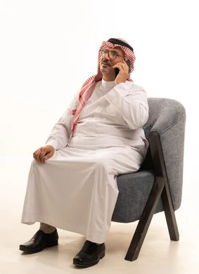 Making a phone call on a mobile device, using modern technologies for remote work, completing daily tasks, a close-up portrait of an elderly Arab Gulf Saudi man wearing traditional attire and a ghutrah holding a mobile phone, accomplishing tasks online through technology, white background.