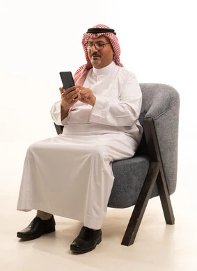 Browsing social media on mobile, using modern technologies for remote work, completing daily tasks, a close-up portrait of an elderly Arab Gulf Saudi man wearing traditional attire and a ghutrah holding a mobile phone, accomplishing tasks online through technology, white background.