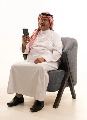 انهاء المهام اليومية، تصفح مواقع التواصل الاجتماعي على المحمول، استخدام التقنيات الحديثة في العمل عن بعد، بورتريه مقرب لرجل كبير السن عربي خليجي سعودي يرتدي الزي التقليدي والشماغ يمسك الهاتف المحمول، إنجاز المهام اونلاين من خلال التكنولوجيا، خلفية بيضاء .