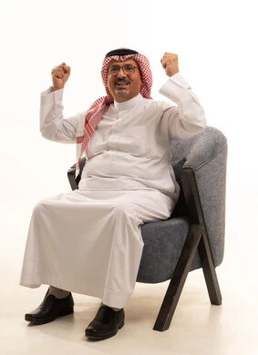 Strength, challenge, and enthusiasm, the concept of achievement and reaching goals, a portrait of an elderly Arab Gulf Saudi man wearing a white thobe and a ghutrah sitting on a comfortable sofa raising his fists upwards in gestures of encouragement, expressions of joy and happiness, white background.