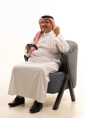 ايماءات الانتصار والفوز، التفاعل والاندماج مع ألعاب البلايستيشن، تقنيات الكترونية حديثة ترفيهيه, بورتريه مقرب لرجل عربي سعودي خليجي كبير بالسن يرتدي الشماغ والثوب التقليدي يجلس على كرسي مريح يحمل بيده عصا التحكم يقوم باللعب في ألعاب الفيديو، خلفية بيضاء