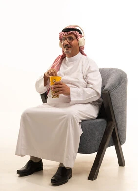 الترفيه عن النفس وقضاء وقت ممتع، إحضار السناكات اللذيذة والاستعداد لمشاهدة الافلام، الاستماع الى موسيقى، بورتريه  لرجل كبير مسن عربي خليجي سعودي مبتسم  يرتدي الزي التقليدي و الشماغ يحمل بيده علبة الفوشار، إيماءات السعادة والمتعة، خلفية بيضاء