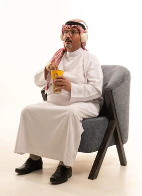 Bringing delicious snacks and getting ready to watch movies, entertaining oneself and having a good time, listening to music, a portrait of an elderly Arab Gulf Saudi man smiling, wearing traditional attire and a ghutrah, holding a box of popcorn in his hand, gestures of happiness and enjoyment, white background.