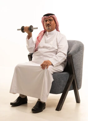 Lifting heavy weights is a physical activity to maintain physical fitness, build and strengthen body muscles. A portrait of an elderly Arab man, a Gulf Saudi, wearing traditional attire and a headscarf, sitting on a sofa holding weights in his hand, showing interest in fitness and physical health, with a white background.