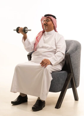 Physical activity to maintain physical fitness, lifting heavy weights, building and strengthening body muscles, a portrait of an elderly Arab Gulf Saudi man wearing traditional attire and a ghutrah sitting on a sofa holding weights in his hand, focusing on fitness and physical health, white background.