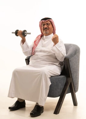 بناء عضلات الجسم وتقويتها، رفع الأوزان الحديدية الثقيلة، نشاط بدني للمحافظة على اللياقة الجسدية، بورتريه لرجل عربي كبير مسن خليجي سعودي يرتدي الثوب التقليدي والشماغ يجلس على الاريكة يحمل بيده الأثقال، الاهتمام باللياقة والصحة الجسدية، خلفية بيضاء
