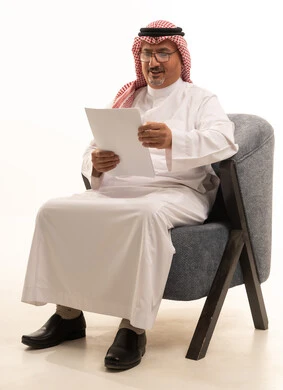 قراءة الملاحظات المدونة من ورقة بيضاء, أدوات تدوين المهام اليومية، تسجيل الملاحظات والمعلومات المهمة,   بورتريه لرجل عربي سعودي خليجي يرتدي الشماغ والثوب التقليدي ونظارة طبية يجلس على كرسي مريح يحمل بيده ورقة بيضاء, بإيماءات التركيز والتمعن، خلفية بيضاء