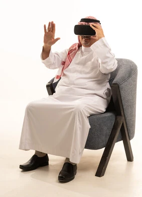 قضاء أوقات ممتعة ومسلية بالألعاب الافتراضية، الاستمتاع بمشاهدة أفلام الواقع الافتراضي، بورتريه لرجل  عربي خليجي سعودي كبير بالسن يرتدي الثوب التقليدي و الشماغ يجلس على كرسي مريح ويرتدي نظارة الواقع الافتراضي ثلاثية الابعاد، التفاعل والاندماج، خلفية بيضاء