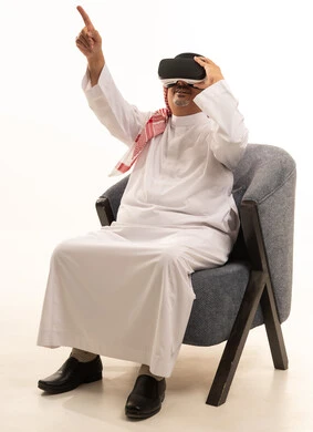 الاشارة إلى شي ما في الأعلى، قضاء أوقات ممتعة ومسلية بالألعاب الافتراضية،  بورتريه لرجل  عربي خليجي سعودي كبير بالسن يرتدي الثوب التقليدي و الشماغ يجلس على كرسي مريح ويرتدي نظارة الواقع الافتراضي ثلاثية الابعاد، التفاعل والاندماج، خلفية بيضاء