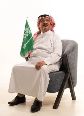 Aging and dignity, holidays and national occasions, celebrating the Saudi National Day, a portrait of an elderly Arab Gulf Saudi man wearing traditional attire and a shemagh sitting on a comfortable chair holding the Kingdom's flag, attention to external appearance, the concept of Arab masculinity, white background.