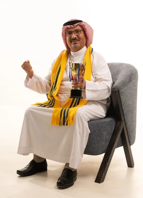 ايماءات الثقة والسرور، الحماس والتفاعل مع المنتخب، الفوز والربح والانتصار، بورتريه لرجل كبير السن عربي خليجي سعودي يرتدي الشماغ والثوب التقليدي يجلس على كرسي مريح يضع الوشاح  يحمل بيده كأس ذهبي، تشجيع الفريق المفضل، أنديه رياضية مختلفة، خلفية بيضاء