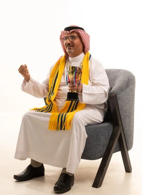 الفوز والحصول على الكأس، الحماس والتفاعل مع المنتخب، الفوز والربح والانتصار، بورتريه لرجل كبير السن عربي خليجي سعودي يرتدي الشماغ والثوب التقليدي يجلس على كرسي مريح يضع الوشاح  يحمل بيده كأس ذهبي، تشجيع الفريق المفضل، أنديه رياضية مختلفة، خلفية بيضاء