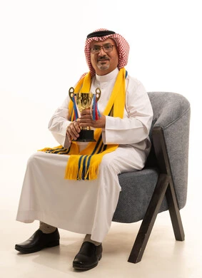 Encouraging the favorite team, winning and obtaining the cup, enthusiasm and interaction with the national team, victory and triumph, a portrait of an elderly Arab Gulf Saudi man wearing a ghutrah and traditional thobe sitting on a comfortable chair with a scarf, holding a golden cup in his hand, different sports clubs, white background.