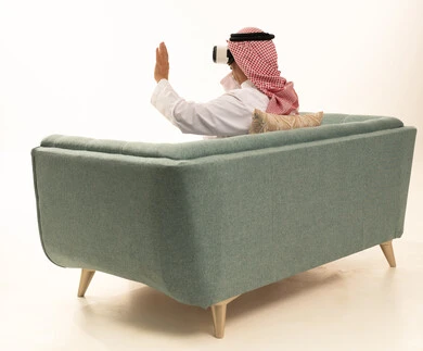 The concept of modern electronic technologies, gestures of interaction and integration with holographic elements, a portrait from behind of an elderly Saudi Arabian Gulf man wearing traditional attire and a ghutrah sitting on a comfortable chair and wearing 3D virtual reality glasses, white background.