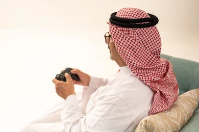 Modern electronic entertainment technologies, spending enjoyable and entertaining times playing, interacting and engaging with PlayStation games, a close-up portrait from behind of an elderly Arab Saudi Gulf man wearing a ghutrah and traditional thobe sitting on a comfortable chair holding a game controller playing video games, white background.