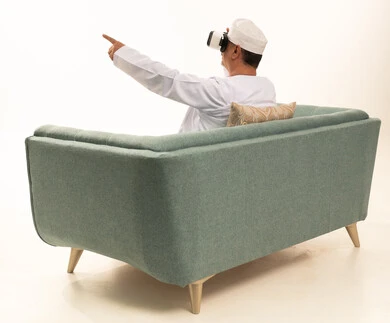 رفع السبابة للاشارة إلى شيء ما، قضاء أوقات ممتعة ومسلية بالألعاب الافتراضية، الاستمتاع بمشاهدة أفلام، بورتريه من الخلف لرجل عربي خليجي عماني كبير بالسن يرتدي الدشداشة والكمة التقليدية يجلس على كرسي مريح ويرتدي نظارة الواقع الافتراضي ثلاثية الابعاد،  خلفية بيضاء