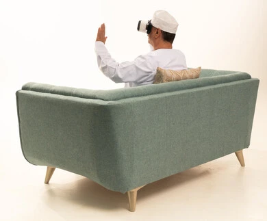 Using the hand to interact and engage with holographic elements, spending enjoyable and entertaining times with virtual games, a portrait from behind of an elderly Arab Gulf Omani man wearing a traditional dishdasha and kumma sitting on a comfortable chair and wearing 3D virtual reality glasses, white background.