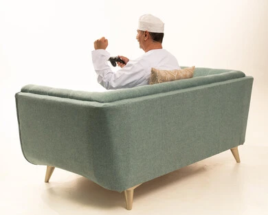 قبضة يد ايماءات فوز وحماس، قضاء وقت ممتع في المنزل، انشطة ترفيهية منزلية، استخدام التكنولوجيا الحديثة في الترفيه، بورتريه من الخلف لرجل مسن عربي خليجي عماني يرتدي الدشداشة والكمة يجلس على الأريكه يمسك بيده عصا التحكم ، المتعة والتسلية والتفاعل، خلفية بيضاء